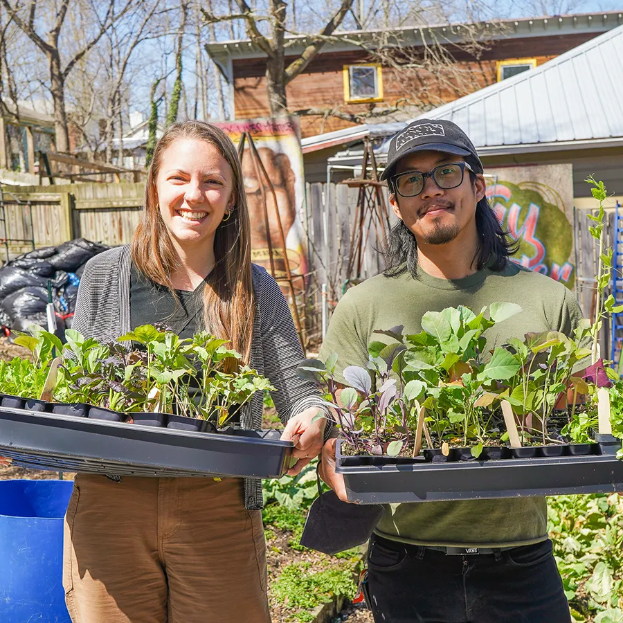 A young woman and young man smile at us holding a flat of of plant starts